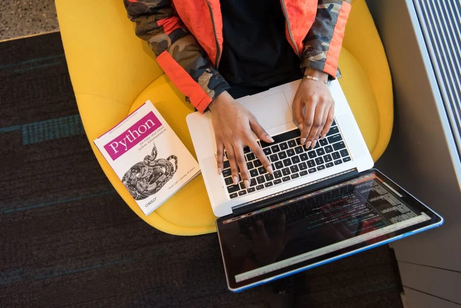 A woman working on a laptop with a camera and smartphone on the desk in a cozy indoor setting.